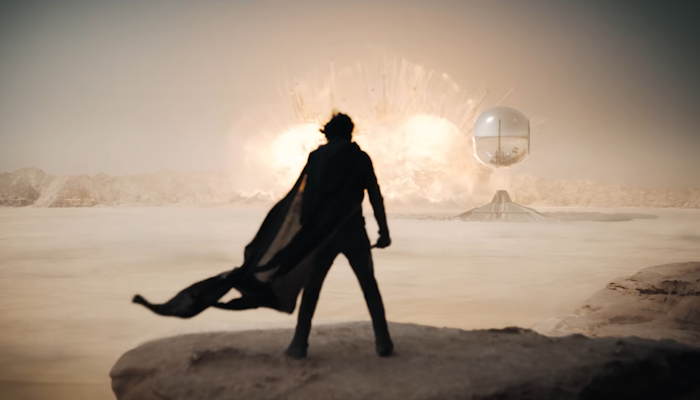 Dune: Part Two, a man stands in the desert, in front of an explosion in the distance.